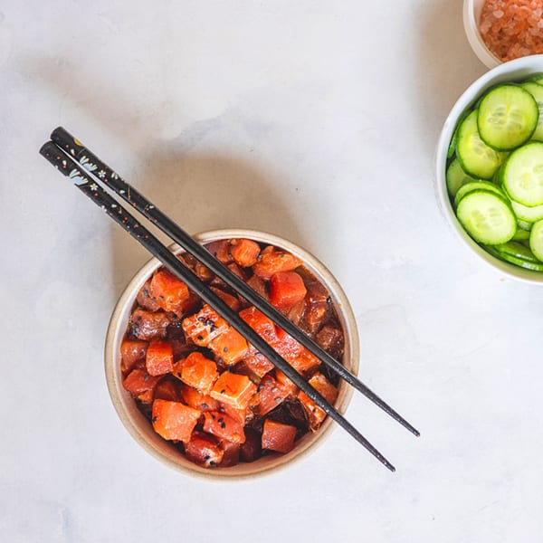 A bowl of diced salmon poke with black sesame seeds and chopsticks, next to sliced cucumbers.