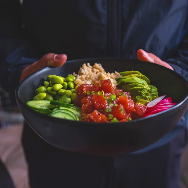 Hands holding a black bowl filled with tuna poke, rice, edamame, avocado, cucumber, and radishes.