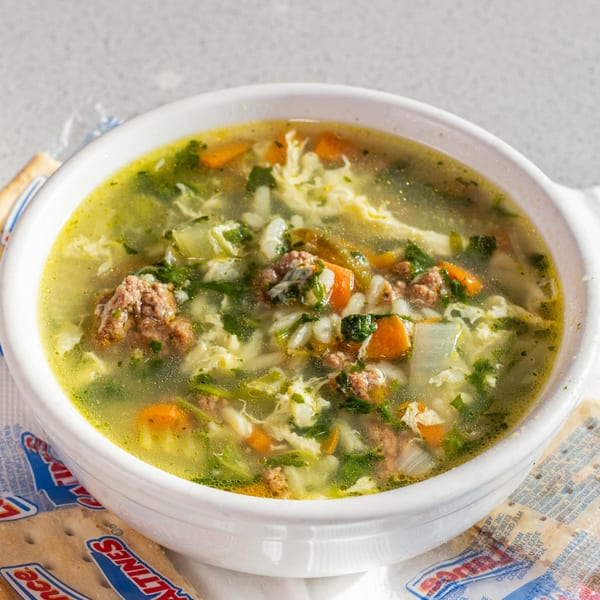 A white bowl of Joe's New York Diner's hearty soup with meatballs, vegetables, and rice, served with saltine crackers.